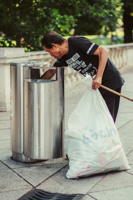 A black wheeled rubbish bin with a white label that reads 'St. John's' is positioned on the pavement next to a curb, partially filled with mixed waste including cardboard and plastic items. The bin is
